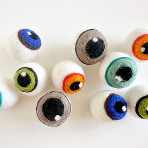 May include: Nine felt eyeballs with different colored irises and pupils. The eyeballs are white with a black outline and are all different colors, including blue, green, orange, and brown.