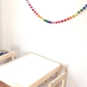 May include: A rainbow felt ball garland hangs on a white wall above a small wooden table and chair set.