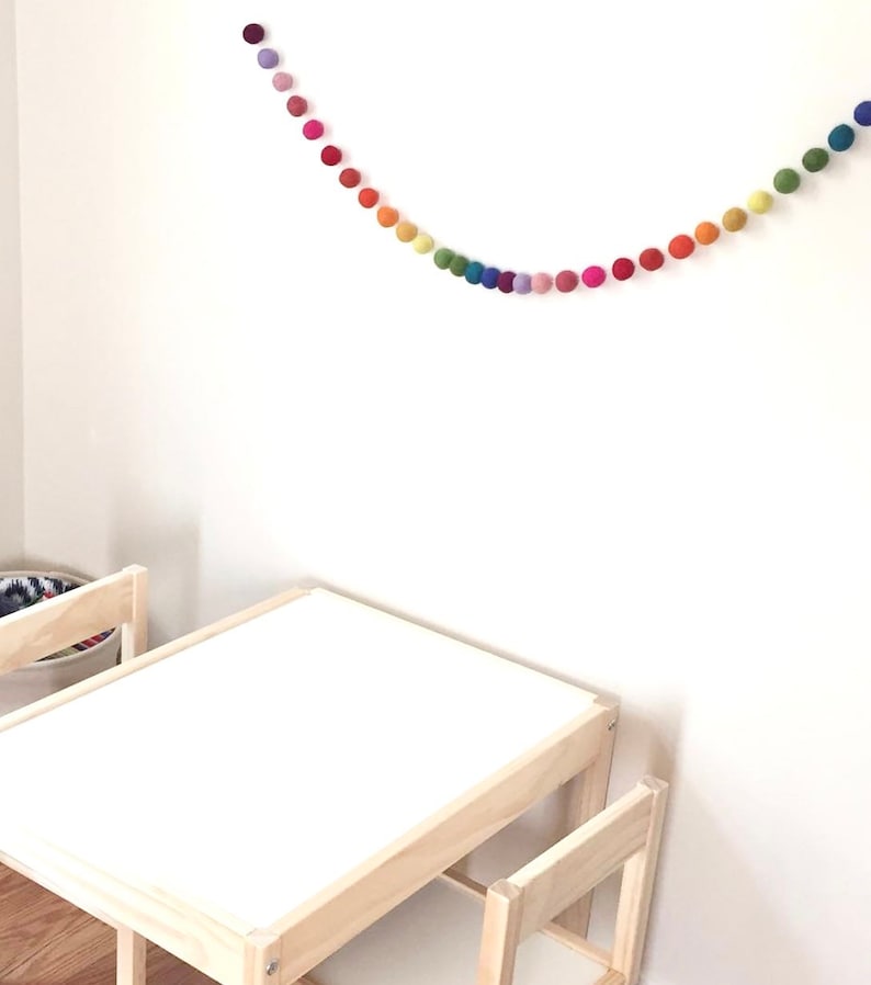 May include: A rainbow felt ball garland hangs on a white wall above a small wooden table and chair set.