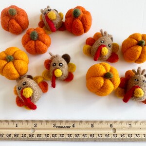 May include: A set of felt pumpkins and turkeys in various shades of orange, brown, and yellow. The felt ornaments are arranged on a white surface with a wooden ruler for scale.