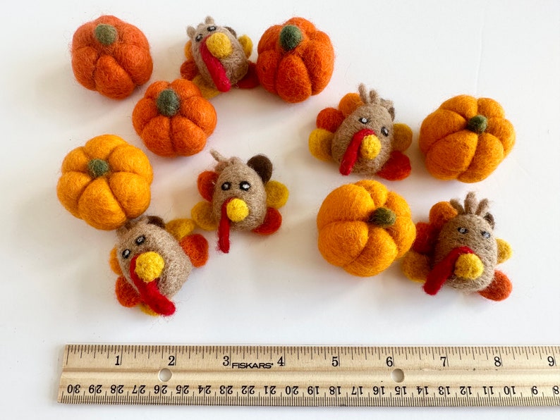 May include: A set of felt pumpkins and turkeys in various shades of orange, brown, and yellow. The felt ornaments are arranged on a white surface with a wooden ruler for scale.