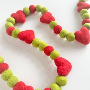 May include: A felt garland featuring red heart shapes and green round beads. The garland is arranged in a curved pattern against a white background. The handmade decoration is suitable for Valentine's Day or other festive occasions.