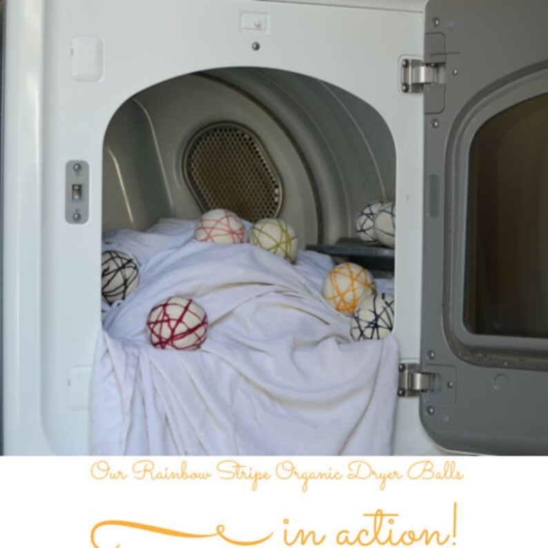 May include: A white dryer door is open, revealing a load of white laundry and several rainbow-striped dryer balls. The balls are various colors, including red, yellow, green, and black. The text at the bottom reads "Our Rainbow Stripe Organic Dryer Balls in action!"