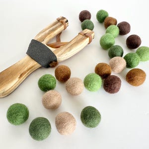 K&ouml;nnte beinhalten: Eine Holzschleuder mit grauem Griff und braunen Gummib&auml;ndern, begleitet von einer Auswahl an bunten Filzkugeln in Gr&uuml;n-, Braun- und Beiget&ouml;nen. Die Anordnung deutet auf ein Spielzeug oder einen Bastelartikel hin.