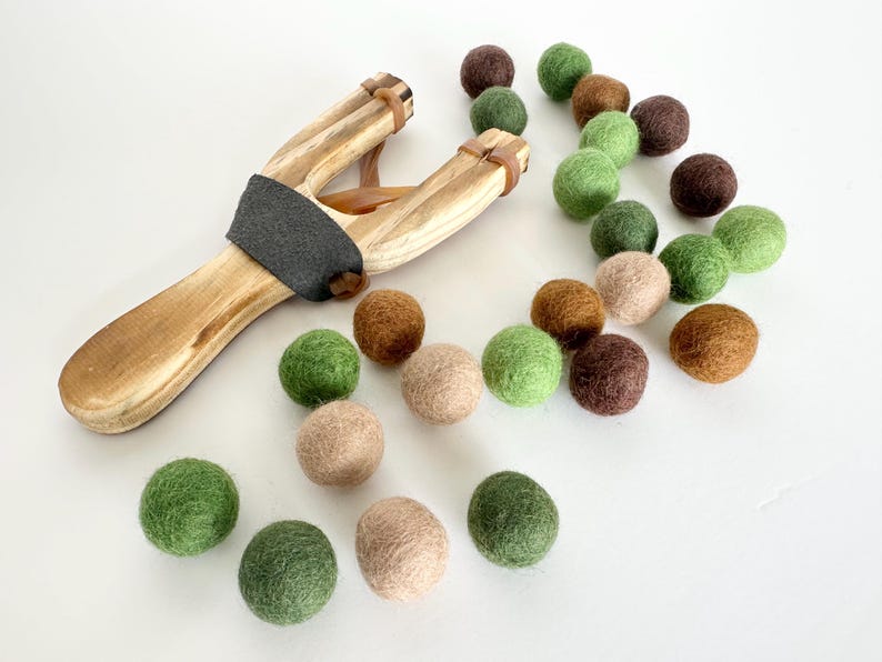 K&ouml;nnte beinhalten: Eine Holzschleuder mit grauem Griff und braunen Gummib&auml;ndern, begleitet von einer Auswahl an bunten Filzkugeln in Gr&uuml;n-, Braun- und Beiget&ouml;nen. Die Anordnung deutet auf ein Spielzeug oder einen Bastelartikel hin.