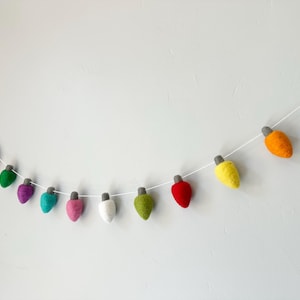 May include: A colorful felt Christmas light garland. The garland features a string of various colored lights, including green, blue, red, purple, teal, pink, white, yellow, and orange. Each light has a gray cap and is strung on a white string.