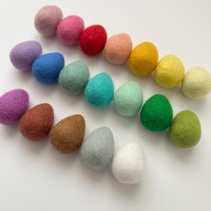 May include: A collection of colorful, egg-shaped felt ornaments in various shades of pink, red, blue, green, yellow, brown, and white. These decorative items are arranged on a white surface, perfect for Easter or spring-themed crafts and decor.