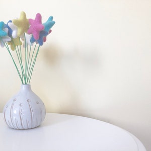 May include: A white ceramic vase with a textured surface and a bunch of felt star-shaped flowers in blue, pink, and yellow colors.