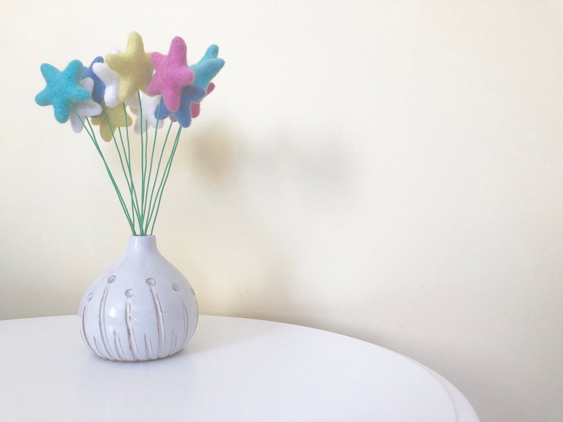 May include: A white ceramic vase with a textured surface and a bunch of felt star-shaped flowers in blue, pink, and yellow colors.