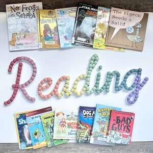 May include: A collection of children's books with colorful covers, arranged on a white surface with a rainbow-colored felt word "Reading" above them.