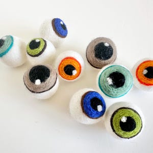May include: Nine felt eye ornaments in various colors, including blue, green, orange, and brown. Each eye has a white sclera and a black pupil.