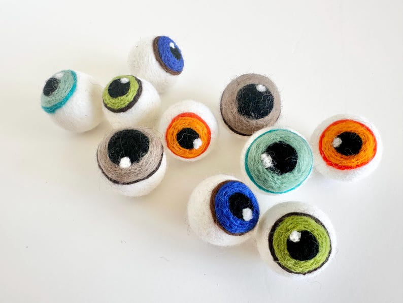 May include: Nine felt eye ornaments in various colors, including blue, green, orange, and brown. Each eye has a white sclera and a black pupil.