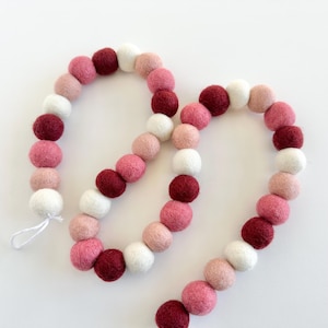 May include: A garland of felt balls in shades of pink, white, and burgundy. The balls are strung together on a white string.