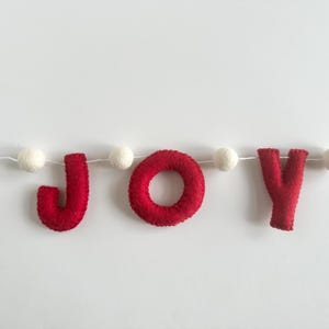 May include: A festive garland featuring the word JOY spelled out in bright red felt letters. The letters are strung together on a thin white cord, interspersed with small, round, white felt balls between each letter to create a decorative banner.
