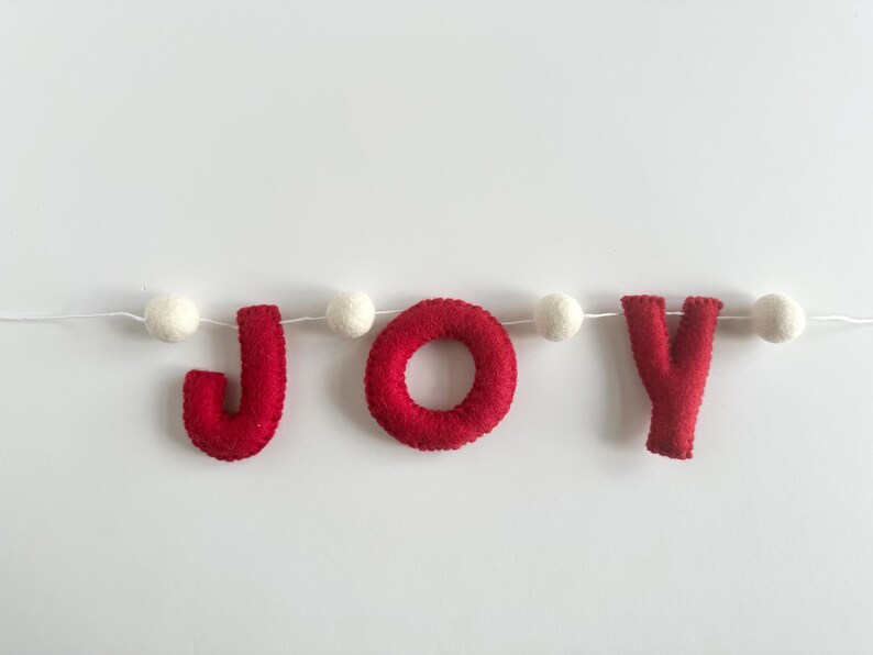 May include: A festive garland featuring the word JOY spelled out in bright red felt letters. The letters are strung together on a thin white cord, interspersed with small, round, white felt balls between each letter to create a decorative banner.