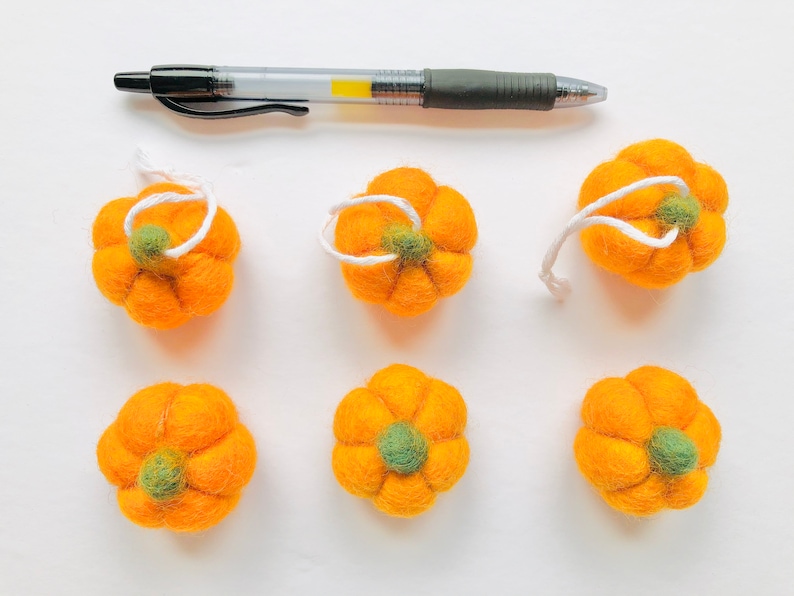 May include: Six felt orange pumpkins with green stems and white string loops. The pumpkins are arranged in two rows of three on a white surface.