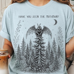 May include: A light blue t-shirt featuring a black ink-style graphic of a forest with a Mothman creature perched atop a central tree. The text "HAVE YOU SEEN THE MOTHMAN?" is printed above the creature, accompanied by stars and a crescent moon.