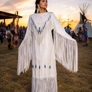 May include: A floor-length white dress featuring long, flowing fringe along the sleeves and hem. The garment is adorned with a V-shaped yoke decorated with blue and black beads, and additional beadwork accents are placed along the lower section.