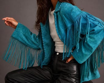 May include: A bright turquoise suede jacket featuring long fringe detailing along the sleeves and front panels, a classic collar, and visible whipstitch trim along the front opening, paired with a white t-shirt and black leather trousers.