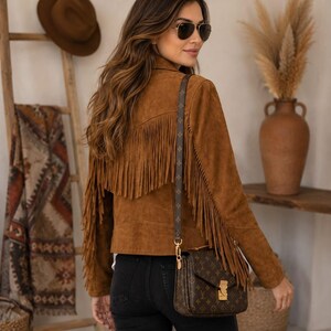 May include: A brown suede jacket featuring long fringe detailing across the back and sleeves, paired with black jeans and a brown monogrammed shoulder bag with gold hardware. The outfit is completed with dark-tinted aviator sunglasses.