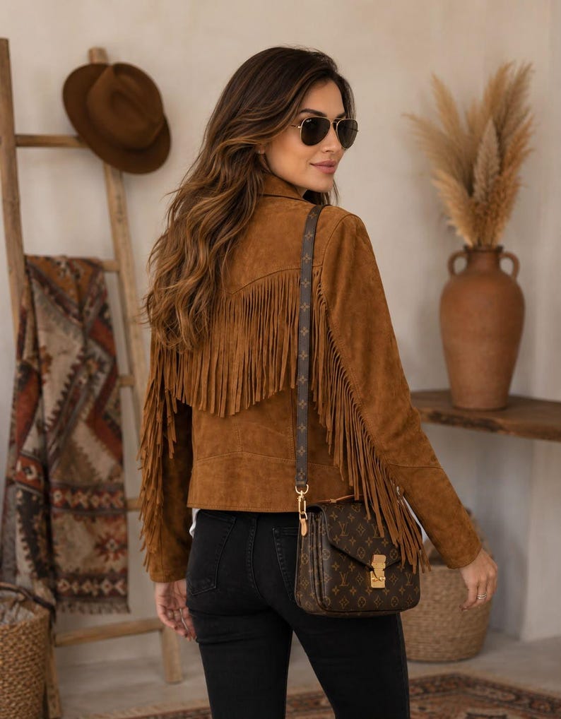 May include: A brown suede jacket featuring long fringe detailing across the back and sleeves, paired with black jeans and a brown monogrammed shoulder bag with gold hardware. The outfit is completed with dark-tinted aviator sunglasses.