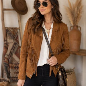 May include: A tan suede jacket featuring long fringe detailing on the front panels, worn over a white button-down shirt and black pants. The jacket has a notched collar and long sleeves, paired with a dark brown patterned crossbody bag.