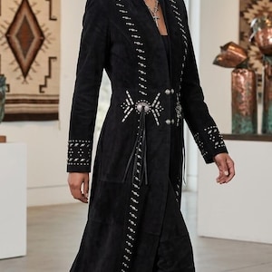 May include: A long black suede coat featuring silver-toned metal studs and decorative concho accents along the front opening, waist, and cuffs. The coat has a high collar, long sleeves, and a structured fit that extends to the lower calf area.