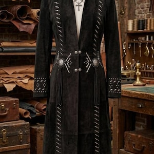 May include: A long black suede coat featuring silver-toned metal stud embellishments arranged in vertical lines along the front and sleeves. The coat includes two circular silver concho accents at the waist, four silver buttons, and long black fringe.