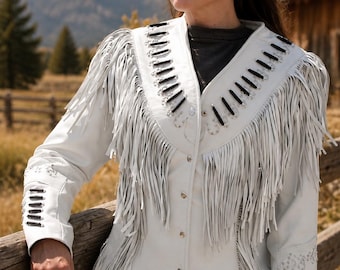 May include: A white leather jacket featuring long fringe detailing along the chest, shoulders, and hem. The jacket includes black cord lacing accents on the lapels and sleeves, silver-toned metal studs, and a front button closure with metallic buttons.