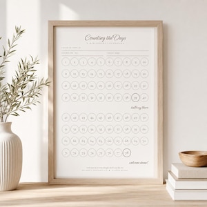 May include: A framed white poster titled Counting the Days with a grid of numbered circles from 1 to 78. The text includes "halfway there" near number 40 and "welcome home!" at the bottom. The frame is light wood with a thin profile.