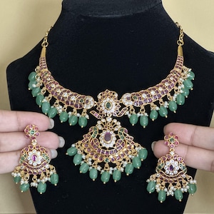 Könnte beinhalten: Ein goldfarbenes Halsketten- und Ohrringset mit filigranen Metallarbeiten, die mit roten und weißen Steinen besetzt sind. Der Schmuck ist mit hängenden hellgrünen ovalen Perlen und kleinen weißen Perlen verziert, was ein verziertes Aussehen erzeugt.