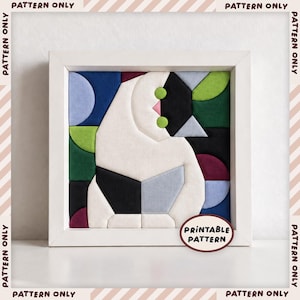 May include: A square framed textile art piece featuring a geometric cat design composed of white, black, blue, green, and burgundy fabric segments. The cat has two bright green circular eyes and a small pink nose. A label reads PRINTABLE PATTERN.