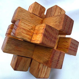 May include: A wooden burr puzzle constructed from multiple interlocking rectangular blocks of light brown oak with a visible wood grain pattern. The puzzle is assembled into a three-dimensional star-like shape with protruding ends.