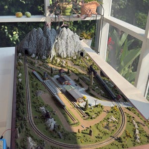 May include: A model train layout on a rectangular wooden base featuring a mountain range with two tunnels, a central station building, a platform, and tracks. A black locomotive with passenger cars travels along the tracks through the landscape.