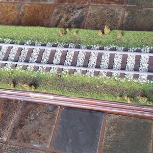May include: A model train diorama featuring a double track set on a bed of gray ballast and green grass. The scene includes a small figurine of a person in a green shirt and blue pants, a tiny white marker post, and a dark wood base finish.