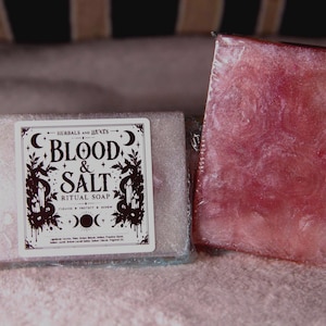 May include: Two rectangular bars of soap wrapped in clear plastic. One bar is light pink with a white label featuring the text "HERBALS AND HEXES BLOOD & SALT RITUAL SOAP" with moon and plant illustrations. The second bar is a deep, translucent red.