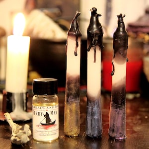 Könnte beinhalten: Drei hohe Stabkerzen mit weißen Kernen und von oben herablaufendem schwarzem Wachs, die an den Basen mit goldenem, blauem und violettem Glitzer versehen sind. Daneben steht eine kleine klare Glasflasche mit dem Etikett Ferry Man Herbal Ritual Oil.