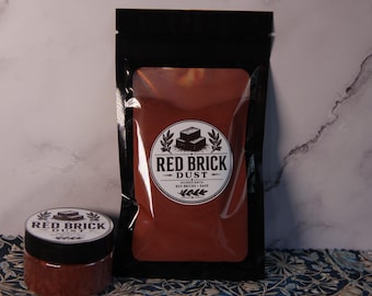 May include: A black stand-up pouch and a small cylindrical jar, both containing a reddish-brown powder. Each container features a circular white label with black text reading RED BRICK DUST and an illustration of two stacked bricks.