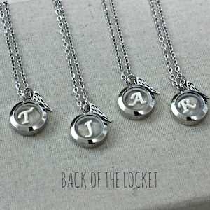 May include: Four silver locket necklaces with angel wings charms. Each locket has a different initial inside: T, J, A, and R. The back of the locket is visible in the image.
