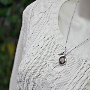 May include: A silver chain necklace with a small, round locket pendant. The locket has a delicate wing design and a clear glass insert. The necklace is worn on a white cable knit sweater.