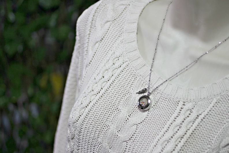 May include: A silver chain necklace with a small, round locket pendant. The locket has a delicate wing design and a clear glass insert. The necklace is worn on a white cable knit sweater.