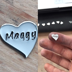 May include: A silver heart-shaped charm with the name "Maggy" engraved on it. The charm is shown next to a smaller version of the charm.