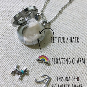 May include: A silver locket necklace with a floating charm of a rainbow. The locket is open and contains a small amount of white pet fur. A silver initial charm is placed behind the fur.