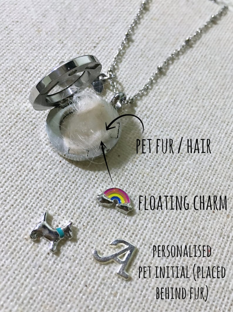 May include: A silver locket necklace with a floating charm of a rainbow. The locket is open and contains a small amount of white pet fur. A silver initial charm is placed behind the fur.