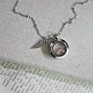 May include: A silver locket necklace with a rainbow charm inside. The locket is attached to a silver chain with a small silver wing charm.
