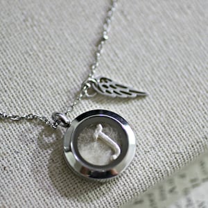May include: A silver necklace with a round locket pendant. The locket has a clear glass window that reveals a small silver letter 'T' inside. The necklace also has a small silver charm in the shape of a wing.