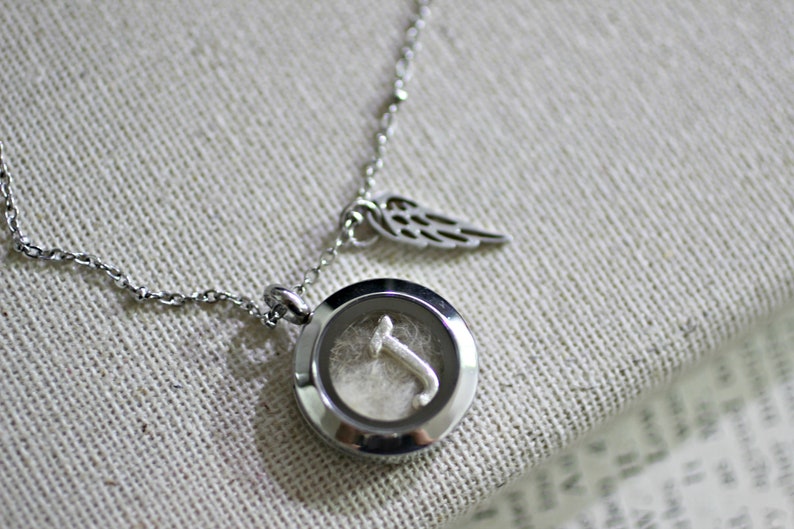 May include: A silver necklace with a round locket pendant. The locket has a clear glass window that reveals a small silver letter 'T' inside. The necklace also has a small silver charm in the shape of a wing.