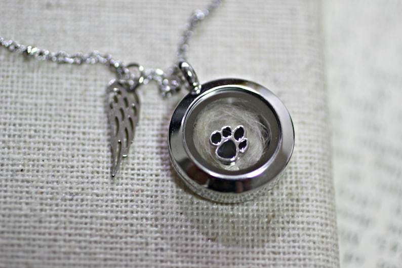 May include: Silver necklace with a round locket featuring a paw print charm inside. The locket is open and contains a small amount of white fur. The necklace also has a small angel wing charm.