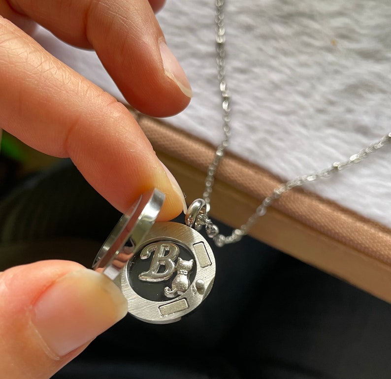 May include: A silver necklace with a round locket pendant. The locket is open, revealing a black background with a silver letter "B" and a small silver cat silhouette.