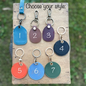 Op de afbeelding: Zeven verschillende leren sleutelhangers met metalen ringen en clips. De sleutelhangers zijn genummerd van 1 tot 7 en zijn in verschillende kleuren, waaronder blauw, bruin, rood, zwart, oranje en groen.